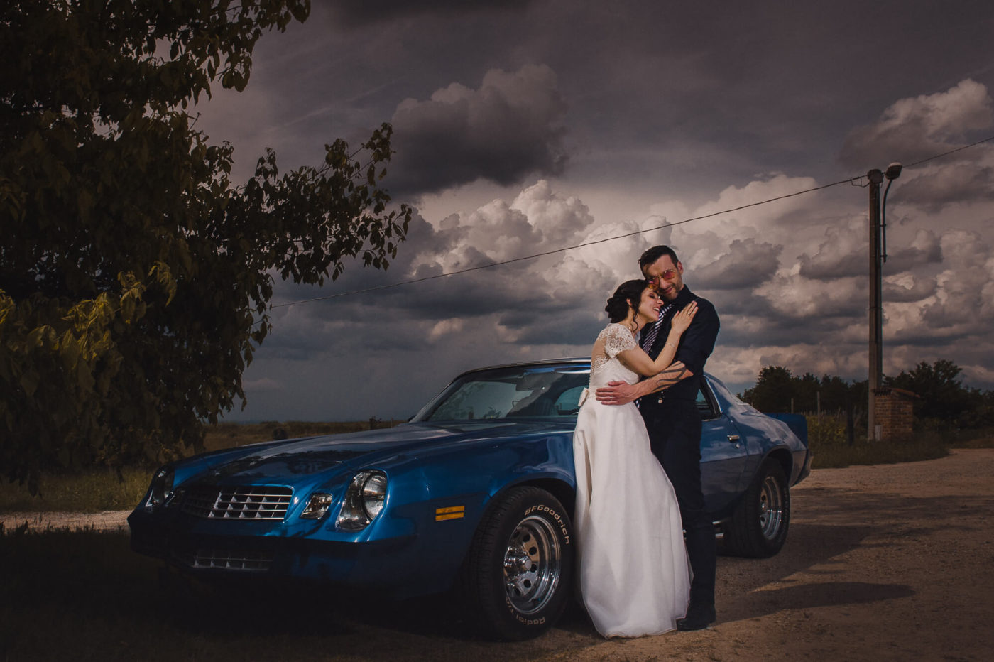 Fotografo di matrimonio Asti - sposi che si abbracciano vicino ad una Camaro blue nel giorno del loro matrimonio sotto un cielo tempestoso